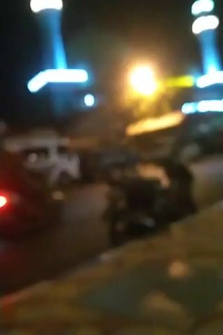 The video begins with a view of a mosque at night, illuminated by blue and white lights on its minarets and dome. A street is visible in front of the mosque, with several vehicles, including a white van, a truck, and a motorcycle. The motorcycle's brake light is illuminated. The camera then pans to the right, showing a sidewalk with a tiled pattern. There are some black trash bags and a pair of sandals on the sidewalk. A person is sitting on the sidewalk, partially visible. Further down the sidewalk, streetlights are visible, and several other people are seated or walking. The video ends with a view of the street and sidewalk, with the mosque and streetlights in the background.
