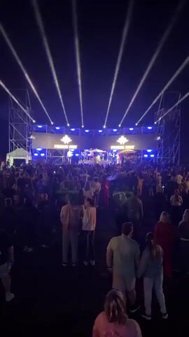 A crowded outdoor event is captured in a vertical video. A stage is set up in the background with a large screen displaying "FRENCH FEST" and numerous blue lights. Several people are on the stage, some appearing to be performers, with bright white spotlights shining upwards from the front. In front of the stage, a large crowd of people is gathered, facing the stage and illuminated by the stage lights. The overall atmosphere appears to be that of a concert or festival.
