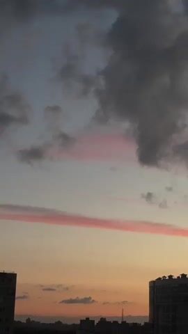 A vertical shot captures a city skyline at dusk. Dark clouds dominate the upper portion of the frame, interspersed with wisps of pink-tinged clouds. A distinct, long, thin pink cloud stretches horizontally across the middle of the sky. Below the clouds, the silhouette of a large, modern building with a distinctive roofline is visible on the right. To the left and further in the background, a darker, less defined city silhouette can be seen. The lower part of the sky transitions from a pale orange near the horizon to a light blue higher up. There are no people or visible actions taking place.