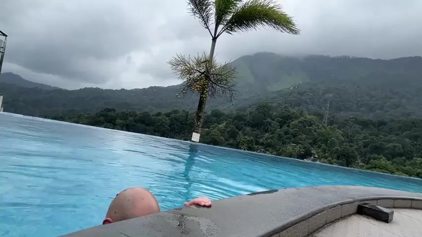 A bald man with stubble is partially submerged in the light blue water of an infinity pool, holding onto its dark gray tiled edge. He looks towards the right with a slight grimace on his face. Behind the pool, a tall palm tree with a slender trunk stands, its leaves swaying slightly. In the distant background, a vast landscape of lush green mountains, partially obscured by mist or clouds, stretches under an overcast sky.