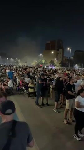 This media file shows an outdoor gathering at night, likely a festival or concert, in l'Hospitalet de Llobregat, Spain. A large crowd of people are milling about, with many seated at tables and chairs. Some people are standing and talking, while others appear to be enjoying the atmosphere. In the background, there are buildings, streetlights, and a stage with lighting equipment. Smoke or haze is visible in the air, suggesting a performance or some sort of fog machine. The overall mood appears to be festive and energetic. The time of day is clearly night, as indicated by the darkness and artificial lighting. The weather appears to be pleasant, with no visible rain or wind.

There is no visible text in the media file.