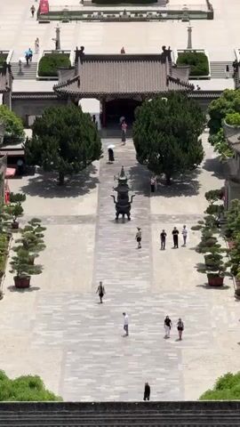 The video shows an aerial view of a long, symmetrical city street or plaza, starting from a vantage point above what appears to be a traditional Chinese building with a tiled roof. The street is lined with trees, interspersed with monuments or statues. People are walking along the street and within the plaza in front of the building. A few cars can be seen on the cross streets. In the background, there are modern high-rise buildings suggesting an urban setting. The overall color palette is dominated by the green of the trees, the red of the traditional buildings, and the grey of the pavement. The day appears sunny. The video begins with a view of the plaza near the building and slowly zooms out to reveal the broader city street.