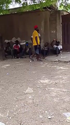 The video shows a group of people gathered outdoors near a building. Several individuals are sitting on the ground, while others are standing. One person is wearing a yellow shirt and a red cap. There is some trash scattered on the ground. The environment appears to be a somewhat rundown urban area. The people seem to be socializing.