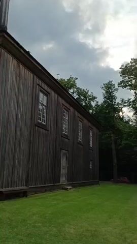 The video shows the exterior of a wooden church with a steeple. The church has wooden plank siding and several windows. There are trees and grass around the church. The sky is cloudy. The camera pans around the building, showing different angles. The sound is of spoken words, but the content is not clear.