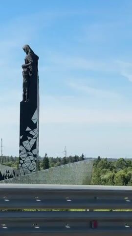 This is a vertical, stabilized video of a dark, tall, obelisk-like monument with a sculpted figure on top. The monument is dark, possibly black, and appears to be made of stone or metal. The sculpted figure at the top depicts a person, possibly a woman, with a child. The lower portion of the monument is decorated with a pattern of shattered or fragmented geometric shapes, which appear to be white or light-colored against the dark background. In the background, there is a clear blue sky with some white clouds. Below the monument, there is a green landscape with trees and some man-made structures, including what appears to be a highway guardrail in the foreground. The overall impression is that of a memorial or monument viewed from a moving vehicle, possibly on a bridge or elevated road.