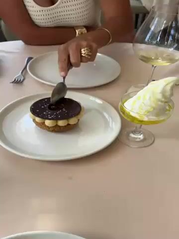 The video shows a person cutting a dessert on a white plate with a fork. The dessert consists of a cookie base topped with white cream, and a dark chocolate disk sprinkled with sea salt. The fork is cutting into the dark chocolate, which has a cutout circle in the center. To the right of the plate is a glass with white ice cream or cream and yellow liquid at the bottom, likely a dessert sauce. Another plate and glass are visible in the background, suggesting a dining setting in a restaurant or cafe. The table has a light pink tablecloth. The person's hand, adorned with gold rings and a bracelet, is visible as they hold the fork.