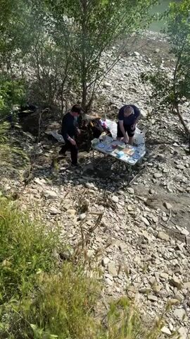 The video shows two men engaged in an outdoor activity on a rocky riverbank. The scene is set under scattered trees and bushes, with a river flowing nearby.

At the beginning of the video, a wide shot captures the two men standing around a portable folding table laden with food items. These include plates, cups, bottles, what appears to be cheese, sliced vegetables, and other picnic provisions. One man, wearing a dark top and shorts, is seen pouring a drink into a cup. The other man, dressed in a dark long-sleeved shirt and trousers, is arranging items on the table. White bags, possibly containing additional supplies or trash, are visible on the ground beside the table. The ground is covered with small to medium-sized rocks and pebbles.

As the video progresses, it zooms in on the table and the men, offering a closer look at the food and their actions. One of the men briefly appears to be opening a bottle. The camera then pans away from the picnic setup, revealing the rocky bank leading down to the greenish water of the river. A small tree with a slender trunk is visible near the water's edge, and a single brick lies on the ground. The sunlight indicates it is daytime, and the overall atmosphere suggests a casual, relaxed outing or picnic by the river. No significant text is visible.