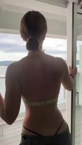 A woman with her hair in a bun, wearing a bikini top, camo pants, and sunglasses is standing on a balcony looking out at the sea and landscape in the distance. She briefly turns to face the camera.
