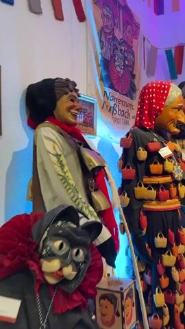 The video shows a display of traditional carnival costumes, likely mannequins. The costumes are elaborate and detailed, featuring masks, hats, and colorful clothing. The masks are particularly striking, with exaggerated features and expressions. Some costumes are adorned with bells, baskets, and other decorative elements. The background of the display includes banners and flags, suggesting a festive or cultural event. There are also informational displays with text and images, which provide context for the costumes. The lighting in the video is dramatic, with spotlights and colored lights creating a moody atmosphere. The overall impression is one of a carefully curated exhibition of cultural heritage.
