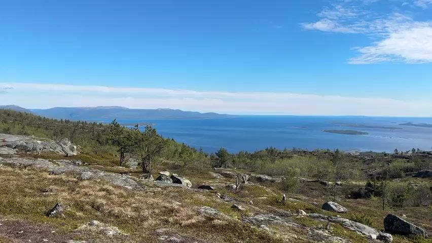 The video shows a scenic landscape, likely in Scandinavia, featuring a rocky terrain with sparse vegetation including small trees and shrubs. Patches of snow remain on the ground. In the distance, a large body of water is visible, dotted with islands and surrounded by hills. The sky transitions from clear blue to partly cloudy with fluffy white clouds. The camera pans slowly across the landscape, showcasing the vastness and natural beauty of the scene.