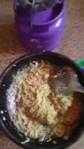 A person is seen stirring a bowl of cooked noodles with a slotted spoon. The noodles appear to be light yellow in color and mixed with some white sauce or broth. The bowl is black and sits on a mottled surface that could be linoleum or tile.  In the background, there are plastic containers of various colors, including purple, green, and blue. The person's hand is visible briefly holding the spoon.  The video is somewhat shaky, and the lighting is adequate.