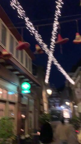 The video begins looking up at a building with a dark red awning and string lights crossing overhead. Three sombreros hang from the awning. The camera pans quickly to the right and then up. The camera points up a street lined with buildings lit by hanging string lights arranged in X patterns. The Domtoren cathedral tower is visible at the end of the street. A man walks in the distance on the street.