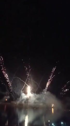 The video captures a nighttime fireworks display over a body of water. Several bursts of fireworks are visible in the dark sky, illuminating the night with streaks of white and red light. Smoke trails are also prominent, dissipating into the darkness. The reflection of the fireworks and lights from the ground can be seen on the surface of the water. In the foreground, a dark silhouette suggests the presence of people or structures observing the display. The overall scene is one of celebration and spectacle, marked by the vibrant explosions of light against the dark canvas of the night sky.