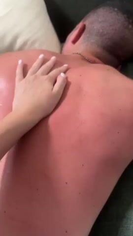 A person's hand with white painted fingernails gently strokes the bare back of another person. The back is flushed red, suggesting a sunburn or rash. The person receiving the touch is lying down, facing away from the camera, and their head is resting on a pillow or cushion. A bottle of what appears to be lotion or oil is visible in the lower right corner of the frame. The scene takes place on a textured green surface, possibly a couch.