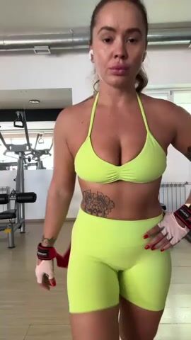 A woman in a neon green workout set, including a halter-style bikini top and high-waisted shorts, stands in a gym. She has tattoos on her arms and abdomen. Her hair is pulled back in a ponytail. She is wearing white and red wrist wraps. She holds red resistance bands in her hands. The background shows gym equipment, including a weight machine and possibly a bench. The room is lit by overhead lights and natural light from windows. The woman looks at the camera. The video appears to be a short clip or boomerang.