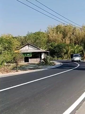 A white pickup truck with a black tarp covering its bed drives down a paved road. The road curves to the right. To the left of the road is a small, weathered building with a lean-to roof extending over the front. The building has a sign that appears to be green and white. The area around the building is covered in dirt and some sparse vegetation. In the background, there are many trees and bamboo stalks. Power lines are visible overhead. The sky is clear and blue.