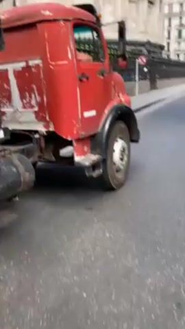 The video begins with a close-up of a faded red truck parked on a city street, with a large building visible in the background. The camera then pans to show a street lined with buildings. As the camera moves down the street, a group of uniformed police officers are seen standing near several parked police motorcycles. Traffic, including cars and a bus, flows down the street. The street is lined with trees and buildings in a city setting.