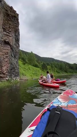 A person is on a paddleboard in the foreground with a blue bag on it. In the middle ground, two red paddleboards are floating on the water. There are three people on these paddleboards, two women and one man. The people appear to be interacting with each other and are facing each other. The woman on the left is holding a paddle. The water is dark and choppy. On the left side of the frame, there is a large, steep, rocky cliff face. Behind the paddleboards and to the right, there is a green, tree-covered hillside. The sky is cloudy and overcast.