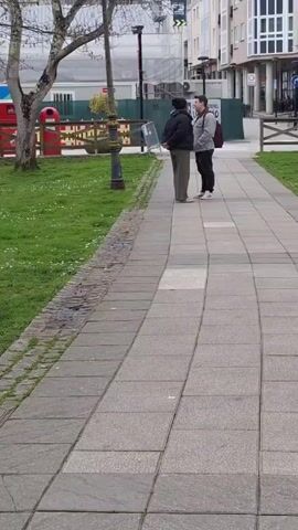 Two people are standing on a paved sidewalk, in what appears to be a small park in a town or city. To the left of the sidewalk is a grassy area with some trees and playground equipment in the background. To the right of the sidewalk are several buildings. The person on the left is wearing dark pants and jacket. The person on the right is wearing a gray jacket, dark pants and carrying a backpack. The person on the left appears to be looking at something on the ground. They are speaking to each other.