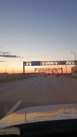 The video shows a view from inside a vehicle driving on a highway during sunset. The sky is a clear blue transitioning to an orange hue near the horizon, indicating the sun is low. There are several overhead gantries spanning the highway, some displaying road signs. Cars are visible driving in the same direction on the highway. The reflection of the overhead structures and the sky is visible on the vehicle's dashboard. The road appears to be a multi-lane highway with streetlights lining the sides.