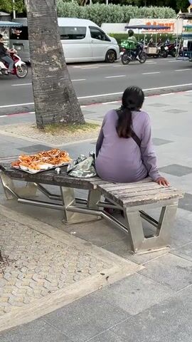 A person is sitting on a wooden bench with metal legs. In front of them on the bench is a tray of food, possibly fried. To the left of the food is a bag. The person is wearing a purple long-sleeved shirt and has their hair tied back. They are facing away from the camera, towards a street with a palm tree in the foreground. Several vehicles, including a white van and motorcycles, are visible on the street. The scene appears to be outdoors, possibly in a tropical or subtropical location, given the palm tree.