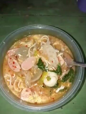 The video shows a close-up, overhead view of a clear plastic bowl filled with a noodle soup. The soup has a reddish-orange broth and contains several ingredients, including sliced hot dogs, large greyish-white pieces that appear to be dumplings or taro cakes, green leafy vegetables, and white noodles. A metal spoon is partially submerged in the soup on the right side of the bowl, suggesting that someone is about to eat or is in the process of eating the meal. The bowl is placed on a green, textured surface, possibly a table. The lighting is dim, and the focus is solely on the food.