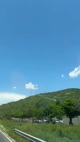 The video shows a two-lane road on a sunny day with scattered clouds.  The road curves slightly as it passes a grassy field and a hillside covered with trees.  A few cars are driving on the road, moving away from the camera.  Power lines are visible on the hillside.  The sky is a clear blue.