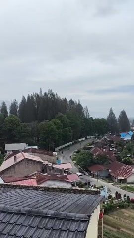 The video provides an aerial view of a town or village under a cloudy sky. In the foreground, a dark grey tiled roof is visible. The scene then transitions to a wider view of houses with red and brown tin roofs, interspersed with green trees and a road. Several houses have blue and white accents. A winding road is visible, with a few motorcycles and cars driving on it. The road is lined with trees and houses. Further in the distance, more houses and buildings can be seen, along with a tall antenna or radio tower. The overall impression is of a moderately populated settlement surrounded by vegetation.