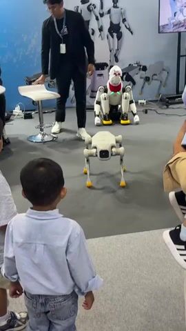 The video shows a group of children and adults watching a demonstration of robots. In the foreground, two young boys are standing and looking at a small, four-legged robot that is white and gray with yellow feet. The robot has a camera on its front. In the background, a man in a black suit is standing and gesturing towards a larger robot that is white with red accents and yellow feet. This robot is also four-legged and appears to be sitting down. Behind the robots is a large display banner with the logo "AGIBOT" and images of several humanoid robots. There are also other people in the background, some sitting at tables. The environment appears to be an indoor exhibition hall with a gray carpeted floor. The overall atmosphere is one of technological display and public interest.