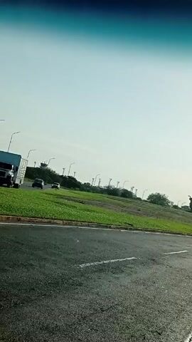 The video shows a view from inside a car driving alongside a grassy median. On the median is a statue of a man on horseback holding a sword. The statue is backlit and appears dark. There are streetlights, trees, and other buildings in the background. The sky is bright.