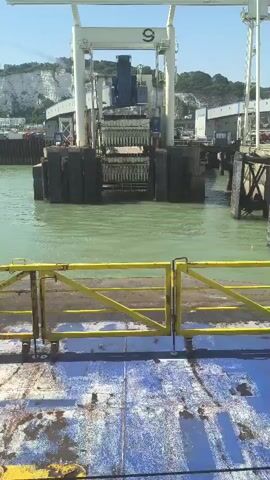 The video shows a view from a ferry looking towards a ferry terminal. In the foreground, a blue deck with some dirt and rust is visible, along with a yellow metal railing. In the midground, green water is present. In the background, a large structure, likely a gantry or lift system, is positioned over the water. This structure has the number "9" prominently displayed on it. Surrounding the gantry are piers and other port infrastructure. White cliffs are visible in the far background, suggesting a coastal location. The sky is clear and blue. The overall scene depicts a busy ferry port environment. There are no visible people or specific actions occurring.