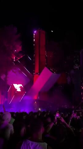 The video shows a nighttime outdoor music festival with a large crowd of people facing a stage. The stage has a distinctive geometric design with red and pink lights illuminating it. The crowd is dense, with many individuals holding up their phones to record the performance. The overall atmosphere appears to be energetic and festive. Trees are visible in the background, and a crescent moon can be seen in the dark sky. The lighting is dynamic, with flashing purple and pink lights sweeping across the stage and the crowd. The sound is not provided, but the visual elements suggest a loud, electronic music performance.