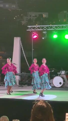 A group of four young women are performing a synchronized dance routine on an outdoor stage at night. They are dressed in matching, vibrant outfits consisting of pink ruffled tops and turquoise skirts with a pattern of red and white dots. Their hair is styled in updos. The stage is illuminated by stage lights, with green, pink, and blue lights visible. Behind the dancers, musical instruments, including drums, are partially visible. The background shows a dark hillside with distant buildings and lights. The camera is positioned from the perspective of an audience member, with the back of someone's head visible in the foreground. The dancers are in various poses, some with hands on their hips and others with arms extended. The lighting shifts and changes throughout the short clip, casting colorful glows on the stage and the dancers.