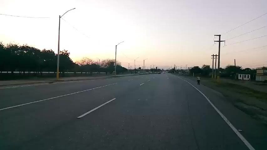 The video shows a view from a vehicle traveling down a four-lane road at dusk or dawn. The road is relatively empty, with only a few other vehicles visible in the distance. The sky is pale with hints of orange from the rising or setting sun. There are streetlights along the side of the road, and power lines overhead. Trees and vegetation border the road, and a lone person is walking on the right-hand shoulder.