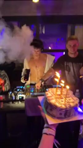 A man in a white shirt stands shirtless at a DJ mixer in a nightclub, performing to music while being enveloped in smoke. Another man with blonde hair stands beside him, also bobbing to the music. In the foreground, a person holds a cake decorated to look like money, with lit candles. Laser lights add to the club atmosphere.