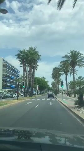 The video shows a first-person perspective of driving on a street lined with palm trees. On the left, there is a multi-story building with balconies and blue awnings. On the right, there is a sidewalk and more palm trees. The road has lane markings and speed limit signs. There are several cars visible on the road, and a few pedestrians on the sidewalk. The sky is partly cloudy. The video appears to be filmed during the day.