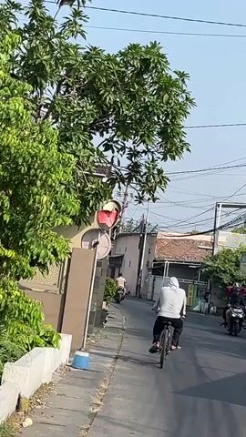A person rides a bicycle down a paved road. The person is wearing a light-colored hooded sweatshirt and dark pants. The bicycle has black tires and a black frame. The road is lined with buildings and trees. There are power lines overhead. In the foreground, there is a traffic mirror attached to a pole. A person on a red motorcycle is visible in the distance.