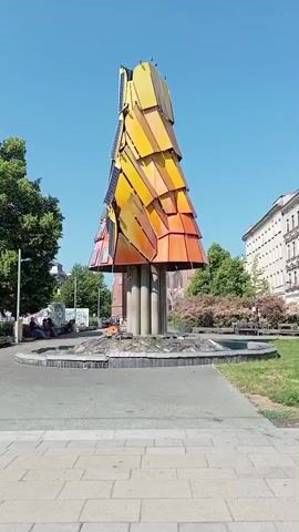 A tall, tree-shaped sculpture made of overlapping, multi-colored metal panels stands in a circular fountain. The sculpture is orange and yellow, with some panels appearing to be solar panels. The fountain has a stone rim and is surrounded by a paved area and a grassy park with trees and buildings in the background. A few people are sitting on a bench or on the ground in the park to the left of the sculpture. The sky is clear and blue.
