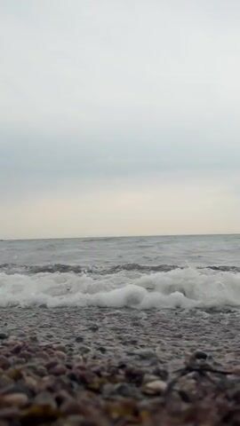 The video shows a close-up, low-angle view of a pebble beach with gentle waves rolling onto the shore. The sky is overcast and appears to be a muted grey. The water is a murky grey-blue, and the waves create white foam as they break. The pebbles on the beach are various shades of brown, grey, and black. The video appears to be a continuous shot with no discernible actions or interactions.
