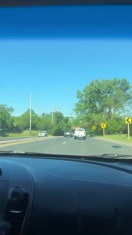 A car is driving down a road with a slight curve. Other vehicles are visible ahead, including a white truck, a silver car, and a yellow convertible. Trees line both sides of the road, and power lines run alongside the trees. The sky is clear and blue. The camera is mounted inside the car, providing a driver's perspective. There is sound of music playing.