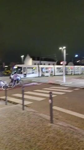 At night, a white tram passes on the street. A person on a bicycle with a trailer pulls across the crosswalk in front of the tram as it clears the intersection. The intersection is at the edge of a town square with streetlamps. The traffic lights at the intersection are red. The sky is dark.