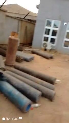 The video shows a person squatting down, picking up a black cloth from the ground. They are wearing a blue top, red pants, and sandals. They appear to be outside, possibly in a yard or work area. In the background, there is a building with white-framed windows, some pipes, and other equipment. The ground is dirt or gravel. The video then pans around to show a wider view of the area, which looks like an industrial or construction site with various barrels, containers, and other objects. It is unclear what the person is doing or what the purpose of the video is.