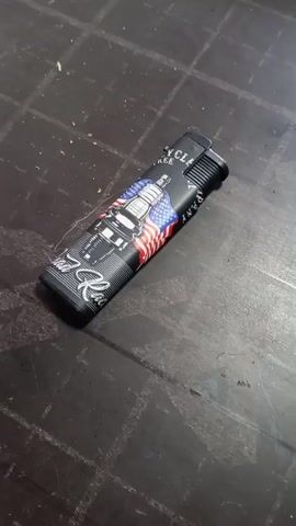 A disposable lighter with a black body and a design featuring a truck and the American flag is placed on a dark, textured surface. A hand enters the frame and quickly moves the lighter out of view. The camera then pans across the surface, revealing a cigarette pack, a tool with red and yellow handles, and scattered debris.