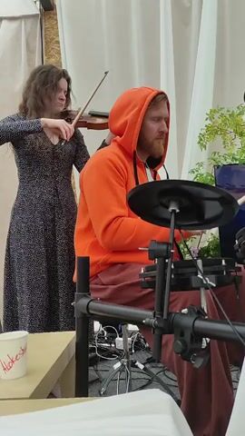 A woman plays the violin while a man in an orange hoodie plays a digital drum set. They are indoors, possibly in a studio or performance space, with white drapes and some greenery in the background. There is a table with a cup on it in the foreground. The lighting is soft and appears to be natural light coming from the right.