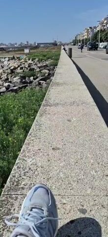 This is a first-person perspective video captured on a sunny day in Vila do Conde, Portugal. The scene features a paved walkway running alongside a rocky coastline. Lush green vegetation with small yellow flowers grows abundantly on the left side of the walkway, leading down to the water.

In the distance, a harbor with numerous sailboats is visible, along with buildings and a lighthouse. The ocean waves crash against the large rocks, creating a dynamic and natural seascape. There are a few people scattered along the rocky area, some appearing to be sunbathing or relaxing. Further down the walkway, other individuals can be seen walking or cycling.

The clear blue sky and bright sunlight suggest it is daytime, likely mid-morning or afternoon. The overall mood is peaceful and relaxing, inviting exploration of the coastal environment. No visible text is present in the video.
