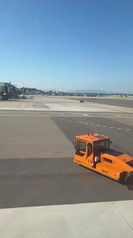 A bright orange tow tractor with the number 41 prominently displayed on its side is positioned on an airport tarmac. The tractor appears to be attached to another piece of equipment at its rear. In the background, an airport terminal with jet bridges is visible. Beyond the terminal, a clear blue sky stretches above a flat landscape dotted with distant buildings and mountains. Several other vehicles, including a white van and a dark-colored vehicle, are present on the tarmac and in the distance. A small airplane can be seen in the sky in the far distance. The scene suggests an active airport environment on a sunny day.