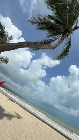 A person's feet are visible in the foreground, resting on a lounge chair with an orange towel. The beach and ocean are in the background, with palm tree fronds in the upper left corner. The sky is blue with white clouds. The ocean has waves and is a greenish-blue color. The sand is light brown.