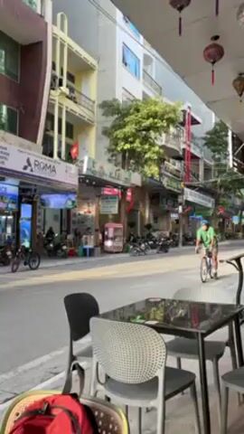 The video shows a cyclist riding down a street in Vietnam. The cyclist is wearing a helmet and a green shirt. He is riding on a paved road alongside several parked motorbikes and cars. Behind the cyclist are several buildings with shopfronts and apartments. The camera is fixed and at an angle capturing the street from under an awning of a cafe. The cafe has a table and chairs in the foreground. A red backpack rests on one of the chairs. There is a Vietnamese flag hanging from one of the buildings. The weather appears sunny. The overall impression is of a typical busy street scene in Vietnam.