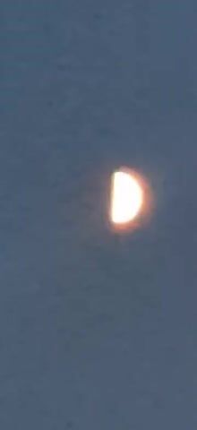 A video shows a half-moon in a dark blue sky. The moon is illuminated on the right side and casts a faint glow around its edges. The sky appears grainy, suggesting it may have been captured with a low-resolution camera or under low-light conditions. There are no other visible celestial bodies or objects in the frame. The video seems to be a static shot with no apparent movement or changes in the scene.