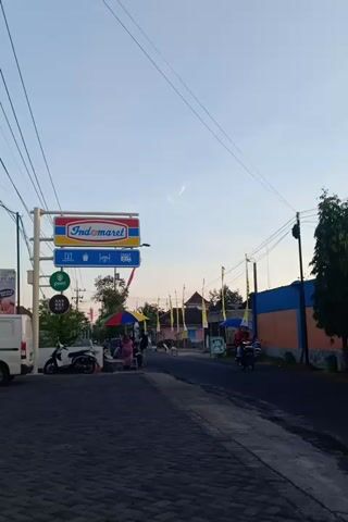 The video shows a street scene with a sign for Indomaret, a convenience store. There are several motorcycles on the street, and some people are visible. The sky is overcast. The video is filmed vertically.